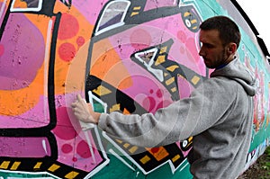 A young guy in a gray hoodie paints graffiti in pink and green c