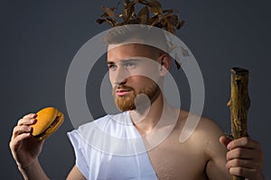 A young guy in the guise of an Olympic god in a laurel vignette with a staff, holds a burgur in his hand, looking at him. Studio