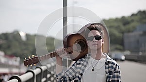 A young guy with a guitar on his shoulder is walking around the city in a shirt and sunglasses. A street musician in