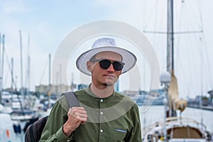 Young guy in a hat and in sunglasses stands in the port of Barcelona at of masts of ships background, Barcelona, Spain.