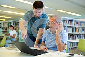 Young guy helping older man in laptop interface in library