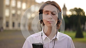 A young guy holds a mobile phone in his hand and listens to music on headphones, standing outdoors in the institute park