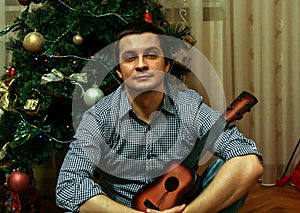 A young guy holds toy guitar sitting on background of New Year tree, blur and grain effect.