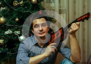 A young guy holds toy guitar sitting on background of New Year tree, blur and grain effect.