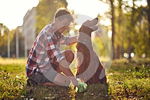 A young guy is kneeling on the grass in the park and collecting the poop of his dog. Friendship, walk, pets