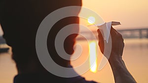 Young guy launch paper plane against the sea during sunset with sun flare and reflections in the water, as in childhood