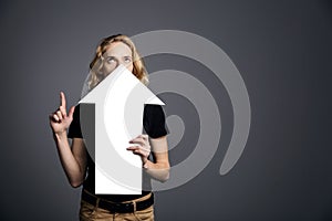 A young guy with long blonde hair is holding in front of his face a white arrow pointing upward on a gray background.