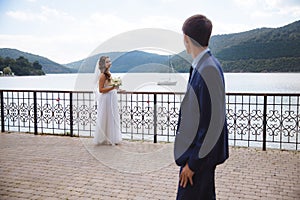 A young guy looks at his bride in a wedding dress against the backdrop of a river along which a boat sails and hills