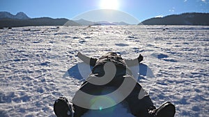 Young guy lying on snow field and making angel on sunny day. Happy man playing and smiling on snowy meadow. Boy enjoying