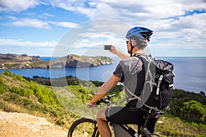 A young guy on a mountain bike trails in Spain and takes a photo on a white phone in the background of the Mediterranean sea of th