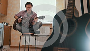 Young guy musician composes music on the guitar and plays in the kitchen, other musical instrument in the foreground,