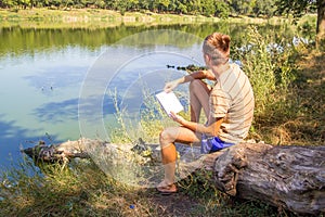 A young guy, with a notebook on the banks of the river, lake, summer thinks records in a notebook thoughts, landscape painting Sun