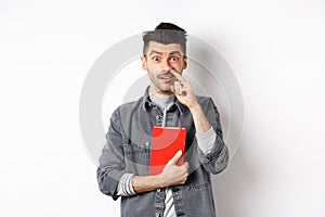 Young guy picking nose and holding book, look stupid at camera, standing against white background