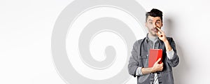 Young guy picking nose and holding book, look stupid at camera, standing against white background