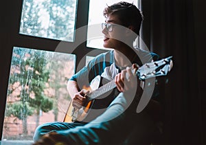 Young guy plaing on guitar and sits near the window