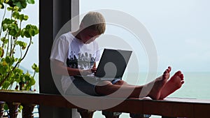 Young guy playing on a laptop, sitting on the terrace. Near the sea and the beach