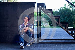 A young guy with purple hair sits on a skateboard in the park on the asphalt