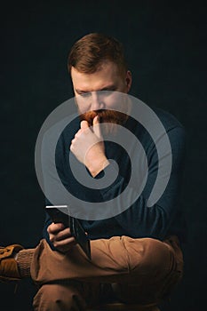 A young guy with a red beard of Irish appearance sits and thinks while reading a phone on a dark background. The concept of the