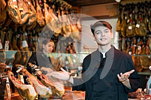 Young guy seller in black jacket selling dry-cured spanish jamon in butcher shop