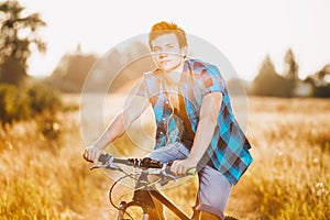 A young guy in a shirt and naked torso sits on a bicycle in headphones and listens to music outdoors, nature, field outside city