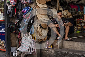 A young guy in shorts, a seller of womens hats and tourist goods in a street shop, plays on his phone.thailand, Phuket