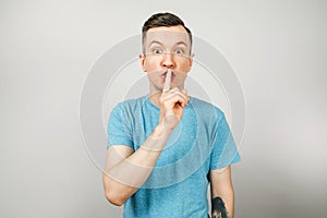 Young guy shut your mouth with finger dressed in a blue t shirt on a light background