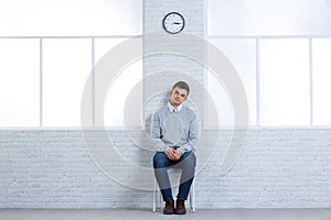 A young guy sits on a chair near the wall and is bored.