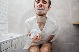 A young guy sits on the toilet and holds toilet paper