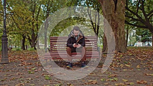 Young guy sitting on a park bench with a phone in his hands.Green tree young man stylishly dressed in the center of the