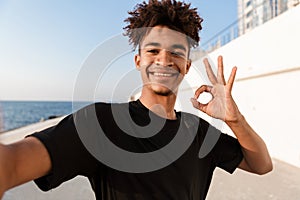 Young guy sportsman standing outdoors on the beach make selfie by camera showing okay gesture