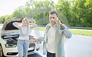 Young guy standing near broken car together with terrified girlfriend, having auto accident, calling breakdown service