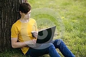 A young guy student sits on the lawn and drinks coffee, holding a laptop in his hands