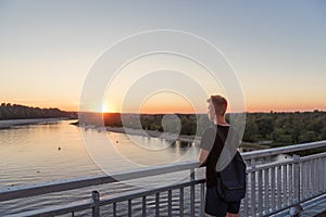 Young guy teenager standing on bridge over river water in lifestyle clothes near steel railing and looking afar at sunset