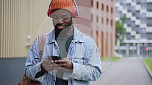 Young guy using device and looking with smile while standing on city street in autumn spbas.