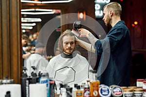 Young guy visiting barbershop. Professional barber drying the hair of his client sitting in armchair in front of the