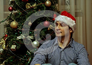 A young guy wearing Santa Claus hat sitting on background of New Year tree, blur and grain effect.