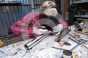 Young guy welder in a checkered red shirt welds a stainless steel pipe for car exhaust using agronomic welding to protect his eyes