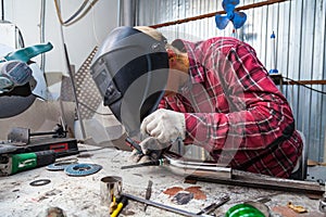 Young guy welder in a checkered red shirt welds a stainless steel pipe for car exhaust using agronomic welding to protect his eyes