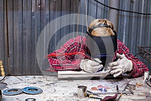 Young guy welder in a checkered red shirt welds a stainless steel pipe for car exhaust using agronomic welding to protect his eyes