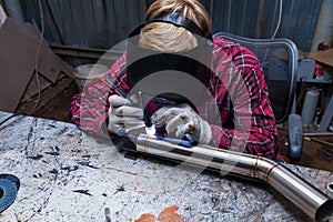 Young guy welder in a checkered red shirt welds a stainless steel pipe for car exhaust using agronomic welding to protect his eyes