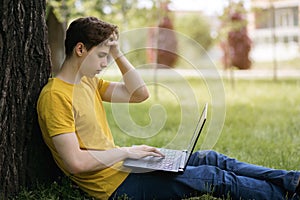 A young guy in a yellow T-shirt sits on the lawn and works hard with a laptop