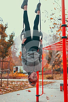 Young gymnast urban woman at playground hanging upside down on rings in park at autumn