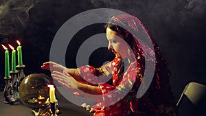 A young gypsy in a red dress at a table by candlelight reads the future on a magical glass ball. The average plan