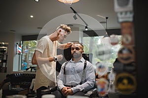 Young hairdresser cutting hair of a male client. A handsome hairstylist serving a client on a blurred barbershop