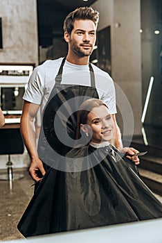 Young hairdresser holding comb with scissors and looking at mirror with beautiful woman