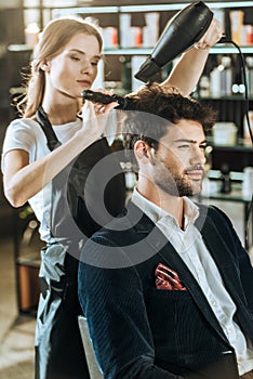 Young hairstylist combing and drying hair to handsome client