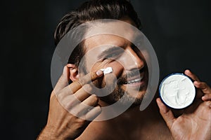 Young half-naked man smiling while applying face cream