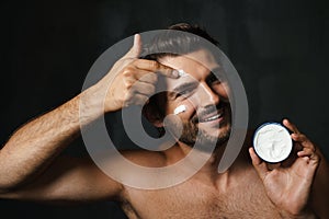 Young half-naked man smiling while applying face cream