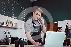 Young handsom male barista wearing apron, talking order on mobile phone and laptop computer while standing behind coffee shop