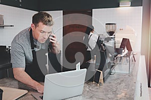 Young handsom male barista wearing apron, talking order on mobile phone and laptop computer while standing behind coffee shop